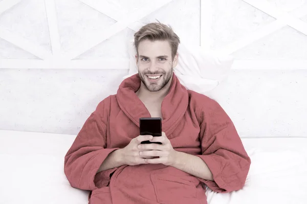 Taşınabilir bir yaşam tarzı. Yatakta mobil cihaz kullanan neşeli bir adam. Elinde cep telefonuyla gülümseyen yakışıklı adam. Mobil iletişim ve evdeki kablosuz teknolojiler — Stok fotoğraf