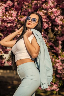 Bahar moda koleksiyonu. Sakura ağacı çiçek açıyor. Bahara karşı tutkulu. Kıyafet dükkanı. Açık havada çok güzel bir kız. Bahar çiçeğinin açtığı süslü güneş gözlüklü kadın. Kiraz çiçekli kız.