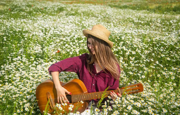 Güzel vakit geçiriyorum. Romantik ruh hali. Bahar doğasının güzelliği. Papatya çiçekleri içinde gitarlı mutlu bir kız. Yaz tatili ve tatil. Güzel bir kadın papatya tarlasında şapka takar. Çayır şarkıcısı
