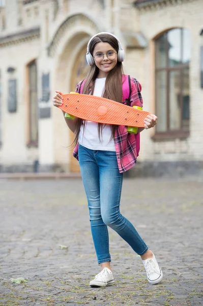 Menina feliz ouvir música em fones de ouvido modernos segurando skate urbano ao ar livre, placa de centavo — Fotografia de Stock