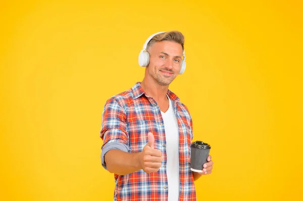 Hombre feliz en los auriculares modernos muestran pulgares hacia arriba gesto de la mano aprobando y promoviendo la taza de café para llevar, placer —  Fotos de Stock