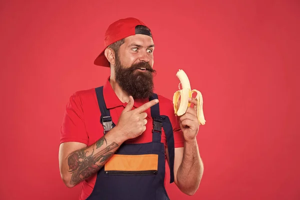 Hungry hipster. Fresh fruit. Living healthy life. Tasty food. Bite banana. Favorite snack. Food court. Lunch time. Bearded man in uniform and cap eating banana. Worker builder mechanic eat banana