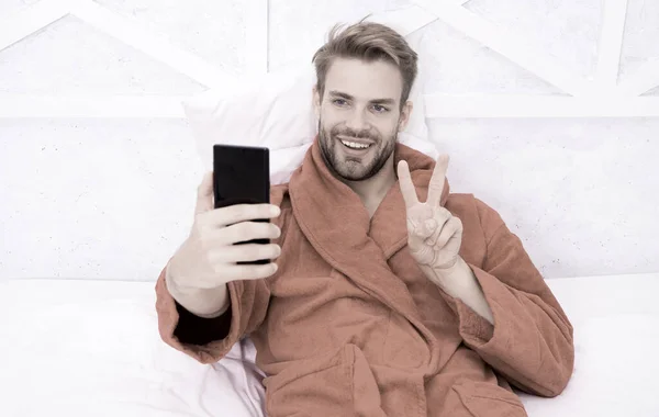Selfie yıldızı. Mutlu adam yatakta akıllı telefonuyla selfie çekiyor. V el hareketiyle cep telefonunda selfie kamerasına gülümseyen yakışıklı adam. Yatak odasından selfie çekmenin keyfini çıkarıyorum. — Stok fotoğraf
