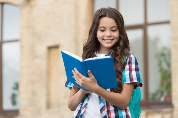 Szczęśliwy dzieciak z długimi falowanymi włosami w stylu casual czytać książkę szkolną na dziedzińcu szkolnym, czytanie — Zdjęcie stockowe