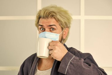 Maskeli adam evde çay ya da kahve içer. Karantina konsepti. Koronavirüsten koru. Adam yüz maskesi takıyor. Virüs damlacıklarla ve yakın temasla bulaşır. Maske takarak koronavirüsten korur.