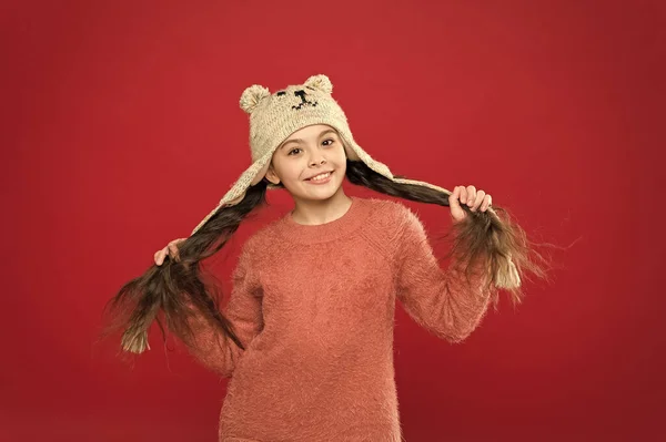 Happiness and joy. Winter outfit. Small child long hair wear hat red background. Cute model enjoy winter style. Adorable small bear. Little kid wear knitted hat. Little girl winter fashion accessory