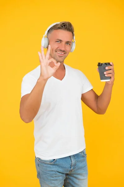 Glücklicher Mann hört Musik, die OK-Handgeste für Kaffee zum Mitnehmen im Einwegbecher gelben Hintergrund zeigt, Coffee to go — Stockfoto