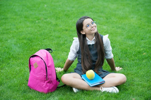 Pensive kid in formal uniform with book and apple healthy school snack relax on green grass, snacking