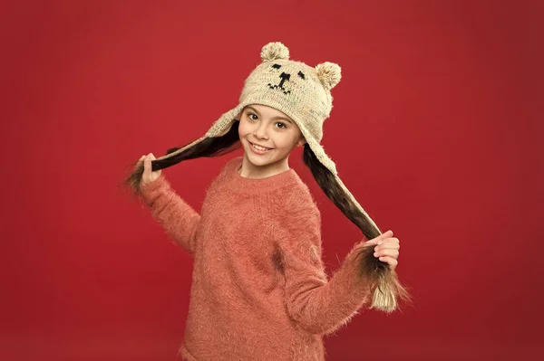 Ευτυχία και χαρά. Το μικρό παιδί φοράει πλεκτό καπέλο. Μικρό κορίτσι αξεσουάρ χειμερινής μόδας. Χειμερινή στολή. Μικρό παιδί μακριά μαλλιά φορούν καπέλο κόκκινο φόντο. Χαριτωμένο μοντέλο απολαμβάνουν το χειμώνα στυλ. Αξιολάτρευτη μικρή αρκούδα — Φωτογραφία Αρχείου