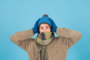 Dışarısı soğuk. Tıraşsız yakışıklı adam mavi arka planda kış aksesuarları takıyor. Alışveriş konsepti. Duygusal bir ifade. Kış soğuğu. Hippi, kış şapkası eşarbı ve eldiven ördü. Sıcak eşarp
