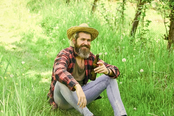 A szalmakalapos farmer sört iszik. érett férfi kertész pihenni zöld fű. Élvezd a tavaszt. A víz eloltja a szomjúságot nyáron. Brutális ranch hipster kockás inget visel. gazdálkodás és mezőgazdaság — Stock Fotó