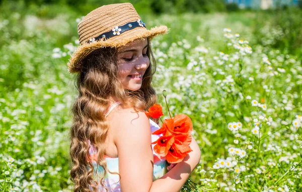 Feliz niña pequeña con aspecto de belleza use sombrero de sol en el pelo largo y rizado rubio sosteniendo flores de amapola en el paisaje natural soleado de verano, salón —  Fotos de Stock
