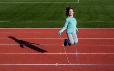 Antrenman rutini. Spor giyimli bir çocuk ip atlarken üzerine atlıyor. Çocuk yarış pistinde egzersiz yapıyor. sağlıklı çocukluk. Açık havada egzersiz yapmak için. İp atlama. Genç kız stadyumda ısınıyor.