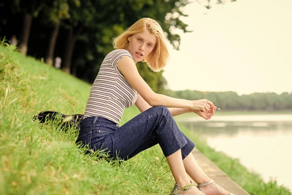 夏休みだ。仕事の後は川沿いでリラックスしている女の子。休暇の夢を見てる女性。リラックスして趣味。自分のために自由な時間を過ごす。少女は川の近くの緑の草の上に座る。休暇の季節 — ストック写真
