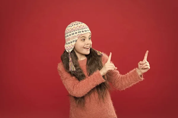 Wskazując w ten sposób. Zimowy strój. Cute model cieszyć się stylem zimowym. Małe dziecko długie włosy nosić kapelusz czerwone tło. Koncepcja czasu zimowego. Mały dzieciak nosi czapkę z dzianiny. Mała dziewczynka zima moda akcesoria — Zdjęcie stockowe