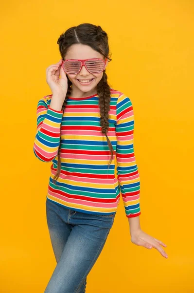 Fröhliche Kind Mädchen mit geflochtenen Haaren tragen bunte Kleidung und Glamour-Party-Brille auf gelbem Hintergrund, Fashionista — Stockfoto