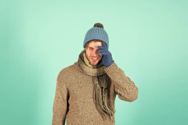 Üzgün hissediyorum. Tıraşsız yakışıklı adam mavi arka planda kış aksesuarları takıyor. Kış sezonu indirimi. Hippi, kış şapkası eşarbı ve eldiven ördü. Alışveriş konsepti. Duygusal bir ifade. Unuttum.