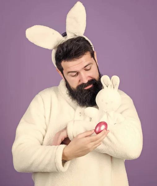 White bunny symbol of easter. Guy with toy and easter egg on violet background. Enjoy tenderness. Spring holiday. Easter when you are parent. Man handsome face wear white bunny ears. Easter bunny