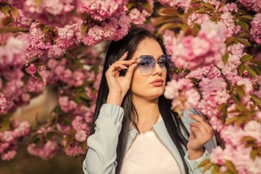 Bahar çiçeğinin açtığı süslü güneş gözlüklü kadın. Kiraz çiçekli kız. Bahar moda koleksiyonu. Sakura ağacı çiçek açıyor. Sakura ona yakışıyor. Muhteşem çiçek ve dişi güzellik. Aksesuar dükkanı.