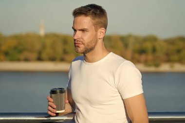 Basit bir yaz eğlencesi. Genç adam nehir kıyısında kahve içmekten hoşlanıyor. Yakışıklı adam sıcak bardağı dışarıda tutuyor. Sabah ayini. Kafein enerjisi. İç ve keyfini çıkar. Kahveden sonra hayatın tadını çıkar. Afiyet olsun.