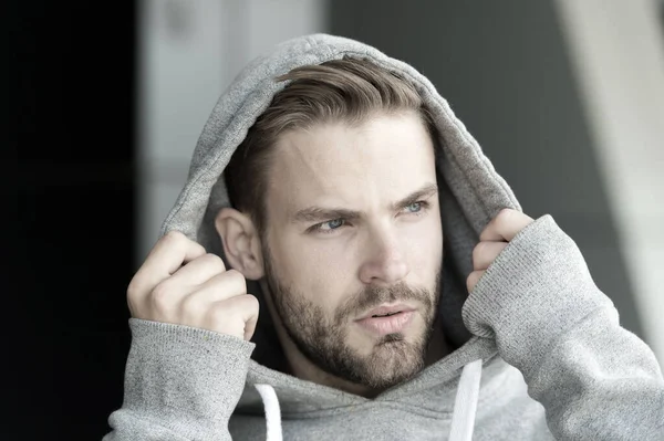 Guy bearded and attractive puts on hood. Masculinity concept. Man with bristle on serious face, urban background, defocused. Man with beard or unshaven guy looks handsome hooded