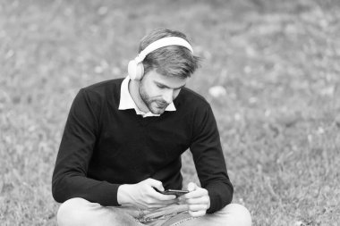 Yeşil çimenlikteki yakışıklı adam cep telefonu mp3 çalarda şarkı değiştiriyor ve dijital çağda kulaklıkla müzik dinliyor.