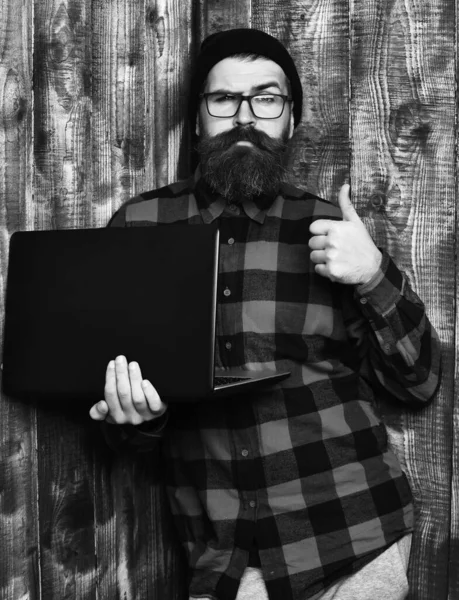 Bearded man, long beard. Brutal caucasian serious unshaven hipster holding laptop in red black checkered shirt with hat and glasses on brown vintage wooden studio background.