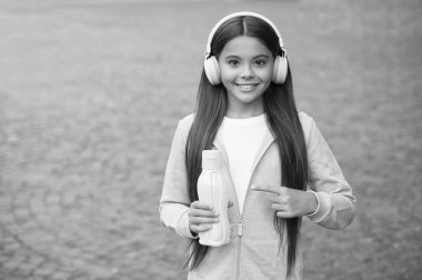 Yüzünde kulaklıkla gülümseyen liseli kız plastik şişeyi işaret ediyor açık havada su içiyor, fotokopi çekiyor, susamış.