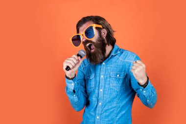 Olgun sakallı adamın uzun saçları var. Parti gözlüğü takıyor ve mikrofonla şarkı söylüyor.