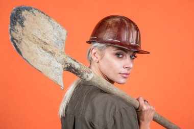 Koruyucu miğfer ve bikini giyen kadın müteahhit turuncu arka planda kürek tutuyor.