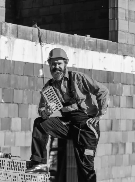 Kendi geleceğini kur. Mutlu sakallı adam tuğla tutuyor. Tuğla işi. Adam yığından tuğla alıyor. Erkek inşaat işçisi tuğla tutuyor. Tamirci adam yeni katman inşa ediyor. — Stok fotoğraf