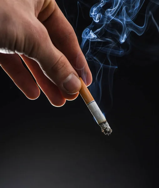 Male hand hold cigarette butt with smoke swirls dark background, copy space, nicotine addiction — 图库照片