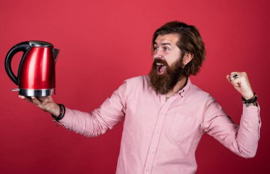 Gömlekli sakallı adam elektrikli su ısıtıcısı tutuyor. Sıradan bir adam modern teknolojiyi günlük hayatta kullanır. Bıyıklı mutlu hipster, kaynar su. Erkek ev idaresi. Çay içme konsepti. yüksek kalite