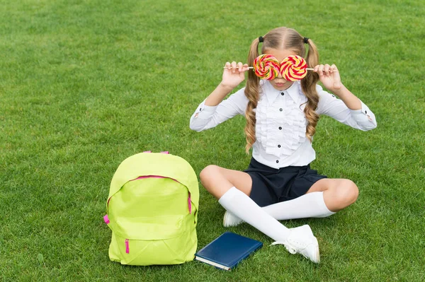 Menina criança no uniforme da escola se divertir cobrindo os olhos com pirulitos na grama verde, dente doce — Fotografia de Stock