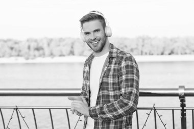 Gündelik gömlekli ve kulaklıklı mutlu bir genç adam açık havada yürürken müzik ve kitap dinliyor.