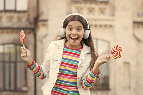 full of happiness. happy schoolgirl in stylish hipster outfit. childhood happiness. kid in digital earphones. small girl listen music with lollipop. back to school. small girl eating sweet candy