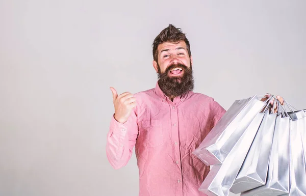 Hipster sur le visage souriant recommande d'acheter. Homme à la barbe et à la moustache porte un tas de sacs à provisions, fond gris. Un type qui magasine pendant la saison des ventes, montrant du doigt une surface vierge. Concept de recommandation — Photo