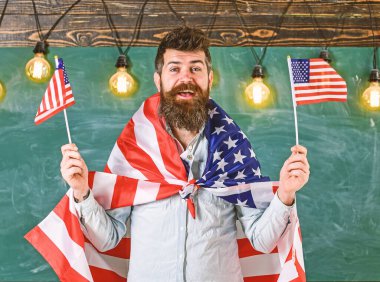 Vatansever eğitim konsepti. Öğrenci değişim programı. Amerikan öğretmeni Amerikan bayraklarıyla el sallıyor. Mutlu yüzlü sakallı ve bıyıklı bir adam ABD bayrakları taşıyor, sınıfta, arka planda karatahta.
