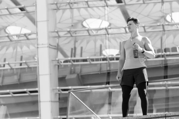 Atleta hombre guapo relajante y beber agua, moderno estadio edificio fondo — Foto de Stock