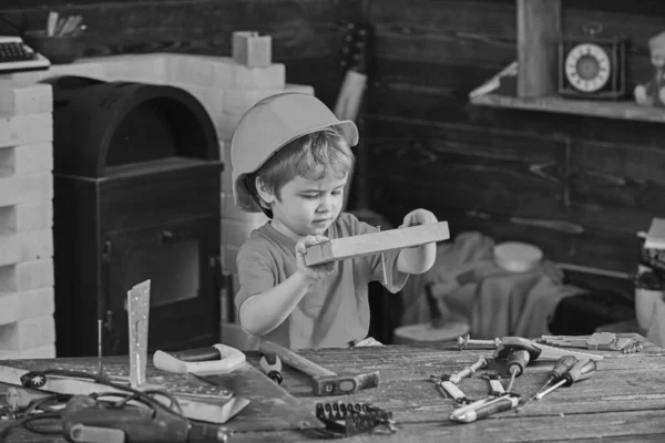 ヘルメットの子供は、ビルダーや修理、修理や手芸としてかわいい演奏。ものづくりのコンセプト。忙しい顔の幼児はワークショップで自宅で演奏します。子供の男の子は木の板に釘を打ち — ストック写真