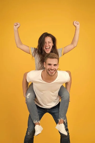 Solid support. Cheerful man piggybacking his girlfriend. Hipster giving sexy woman piggyback ride. Happy couple in love on yellow background. Loving couple smile having fun. Simple happiness