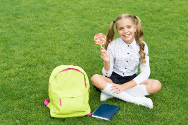 Okul üniformalı mutlu kız, yeşil çimlerin üzerinde oturan tatlı lolipopu tut, şeker.