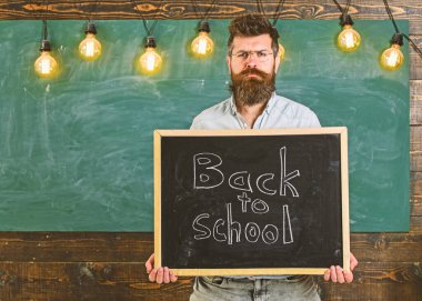 Gözlüklü öğretmen karatahtaya yazı yazmış. Sakallı ve bıyıklı bir adam sıkı yüzlü öğrencileri karşılar, arka planda kara tahta vardır. Okul konseptine dönelim.