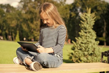 Tecrübe edin. Küçük kız yaz parkında tablet kullanıyor. Modern bilgisayar aygıtı. Dokunmatik ekran arayüzü. Modern teknoloji. İnternette oyun oynuyorum. Video izliyorum. Eğlence ve eğlence. Modern yaşam tarzı