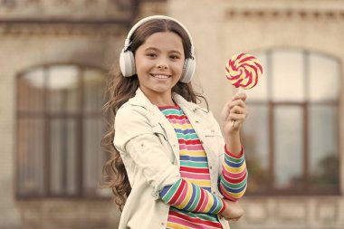 Küçük kız şeker yiyor. Havalı hippi kıyafetleri içinde mutlu bir kız öğrenci. çocukluk mutluluğu. Dijital kulaklıklı çocuk. Küçük kız lolipopla müzik dinliyor. Okula geri dön.