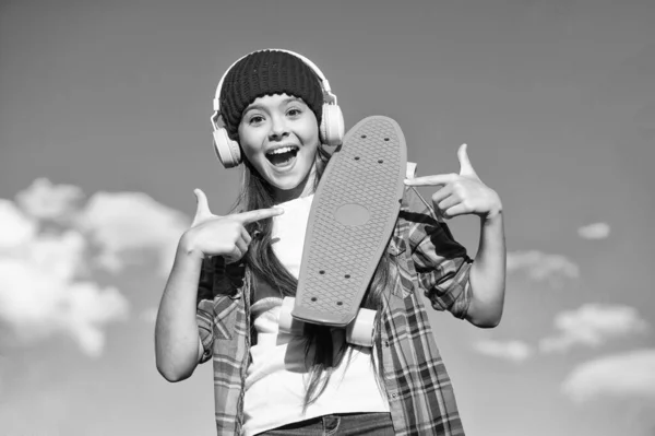 Skateboard you can trust. Happy girl pointing at penny board. Little skater with pointing gesture on blue sky. Pointing for advertising. Marketing and promotion. Good for transportation, copy space