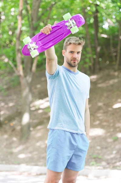 Who want to learn extreme trick. Guy carries penny board ready to ride. Man serious face carries penny board park nature background defocused. Man likes to ride skateboard and sporty lifestyle
