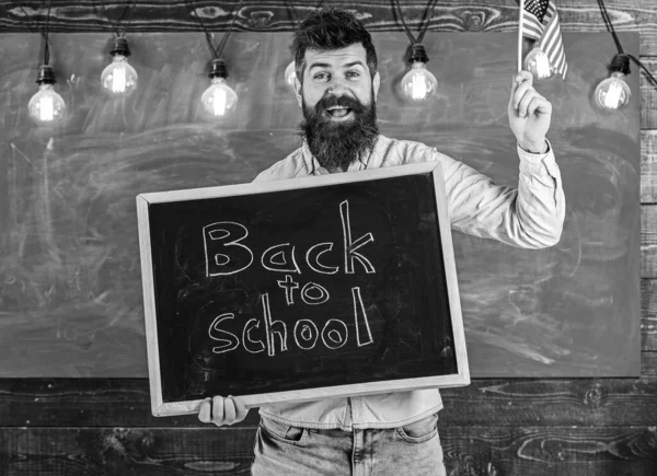 Вчитель тримає дошку з письмовою фразою назад до школи і прапором США. Людина з бородою на усміхненому обличчі вітається в американській школі, на дошці на задньому плані. Концепція американської школи — стокове фото