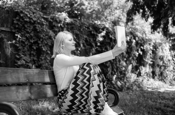 天気の良い日は屋外で読書をするのに忙しい本ワームの女性。文芸評論家。女性は庭で読書の本を集中した。女性はベストセラーについてのレビューを準備します。女の子座ってベンチ本自然背景を読む — ストック写真