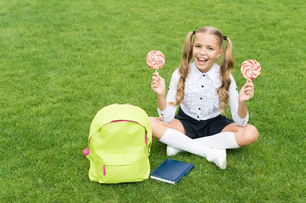 Šťastné dítě ve školní uniformě drží sladké lízátka sedí na zelené trávě, sladkosti — Stock fotografie